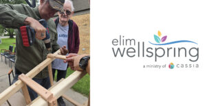 Jerry, a resident at Elim Oasis Adult Day Services, uses a power drill to work on a woodworking project, with a staff member assisting him. Another resident observes in the background, showcasing a hands-on activity that engages participants.